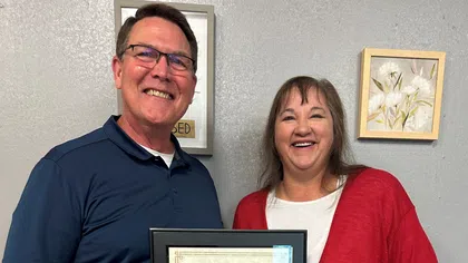 Two smiling adults holding a framed award certificate indoors against a gray wall with floral artwork
