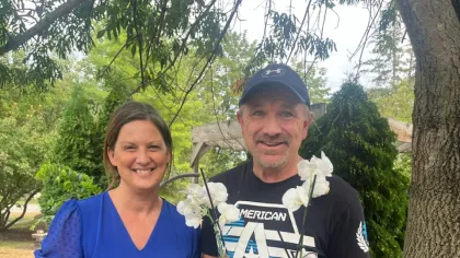 Smiling woman holding award plaque and man with white orchid plant standing outdoors by tree.