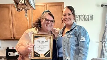 Two smiling women celebrate with a certificate and star-shaped balloons in a cozy home kitchen setting.