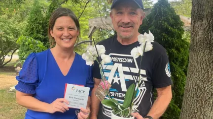 Smiling woman holding award plaque and man holding white orchid plant outdoors in garden setting.