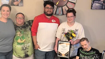 Group of five people smiling indoors, one holding certificate and flowers, others standing or sitting nearby.