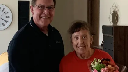 Two smiling people holding an award certificate and a bouquet of flowers indoors, celebrating an achievement.