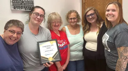 Group of six women smiling indoors, one holding a framed certificate, celebrating an achievement.