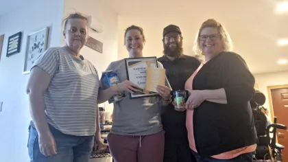 Four smiling adults standing indoors, one holding a certificate and items, casual clothing in a home setting.
