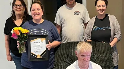 Group photo with a smiling woman holding certificate and flowers, standing behind a seated man in a white shirt.