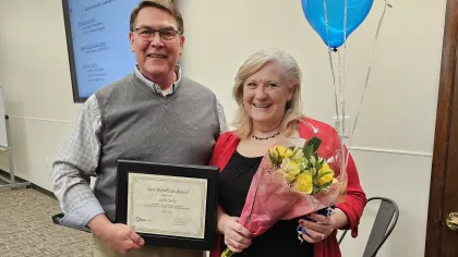 Man holding framed certificate and woman holding bouquet of flowers and balloons in office setting