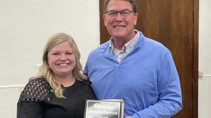 Smiling woman and man holding an award plaque in an indoor office setting with a wooden door background.