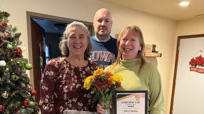 Three adults celebrate with cake, flowers, and a certificate near a decorated Christmas tree indoors.