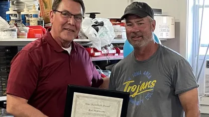 Two men in casual work attire holding a framed award certificate in a workshop with tools and supplies.