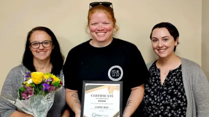 Three smiling women standing together, one holding flowers, another holding a certificate of achievement.