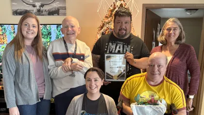 Group of six diverse adults smiling indoors with certificate and gift, in a casual home setting with wooden floor.