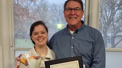 Smiling woman holding bouquet and man holding framed certificate in office with window and balloons
