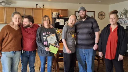 Group of six adults smiling and standing together in a cozy kitchen, one holding a certificate and envelope.