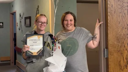 Two women celebrate with a certificate, gift bag, and balloons in an office hallway decorated with vines.