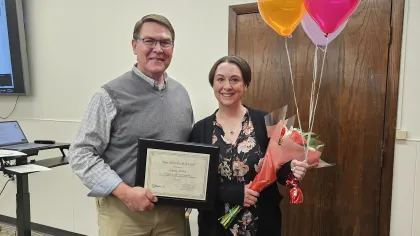 Two people smiling, one holding a certificate, the other holding bouquet and colorful congratulations balloons indoors.