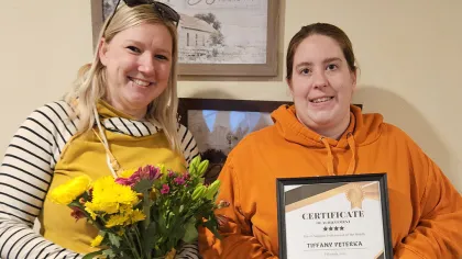 Two women smiling, one holding a bouquet of flowers, the other holding a certificate of achievement indoors.