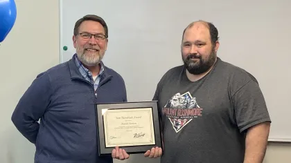 Two men standing side by side holding a framed certificate with a basket of snacks on the table in front