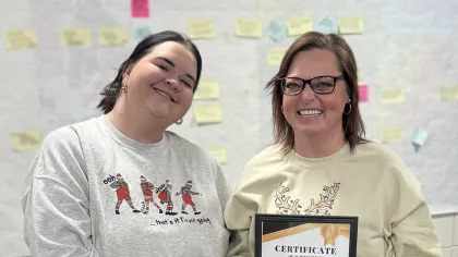 Two women smiling indoors, one holding a certificate of achievement, the other holding a gift bag.