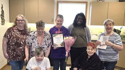 Group of smiling women and seniors, some in wheelchairs, celebrating with flowers and certificates indoors