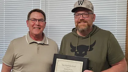 Two men smiling indoors, one holding a framed Tom Thornblade Award certificate, standing in front of window blinds.
