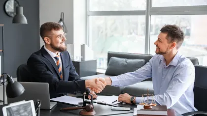 Two business professionals shaking hands in a modern office setting with legal elements present.