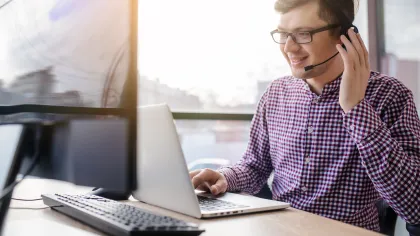 Man in checkered shirt working on laptop, using headset for customer support in bright office space.
