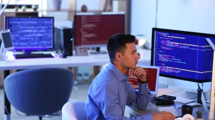 A focused man coding at a computer in a modern tech workspace with multiple screens.