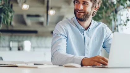 Smiling man working on a laptop in a modern office with plants and bright lighting.