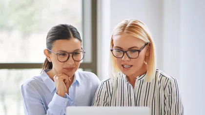 Two women collaborating while looking at a laptop in a modern office setting.