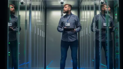 A technician standing in a data center holding a tablet, with reflections in glass panels and server lights.