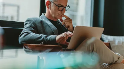 A thoughtful man in glasses using a laptop while sitting in a modern office lounge.
