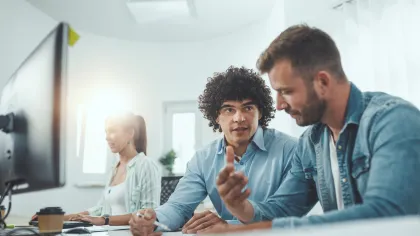 Two men discussing strategies at a modern workspace, with a woman working on a computer in the background.