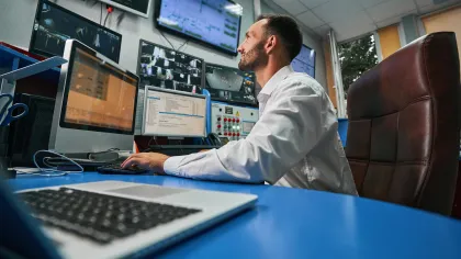A man in a white shirt working at a control desk surrounded by multiple screens.