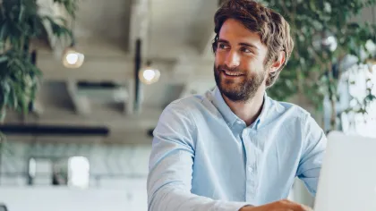 Young professional man smiling while working on a laptop in a modern office setting.