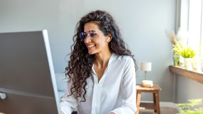 Smiling woman wearing glasses, working on a desktop computer in a bright home office.