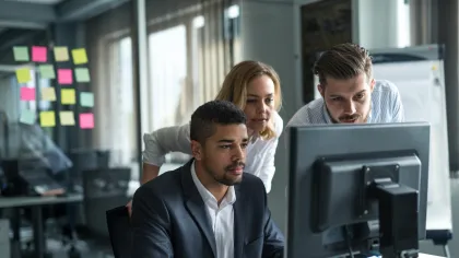 Professionals collaborating in an office, focusing on a computer screen with notes in the background.