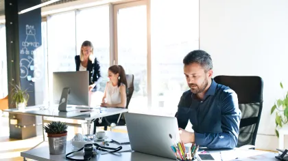 Three professionals working in a bright office space with computers and fresh plants.