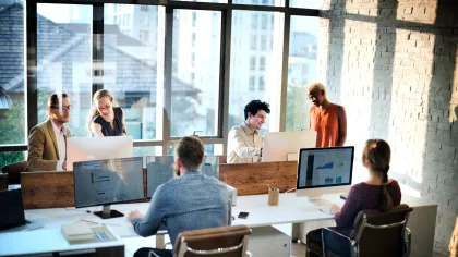 Collaborative office setting with team members working at desks and discussing at large windows.