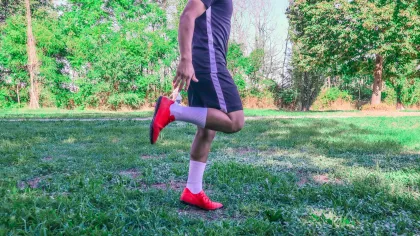 Man in sportswear and red shoes stretching his leg in a green park with trees and grass in the background