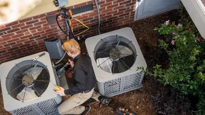Technician repairing two air conditioning units outside a brick and yellow siding house, with tools on the ground nearby.