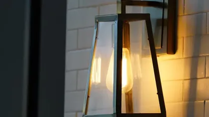 Modern wall-mounted lantern light with clear glass and warm glowing bulb on white tiled wall.