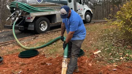 Worker handling green hoses connected to Patriot Plumbing septic service trucks in outdoor setting with trees.