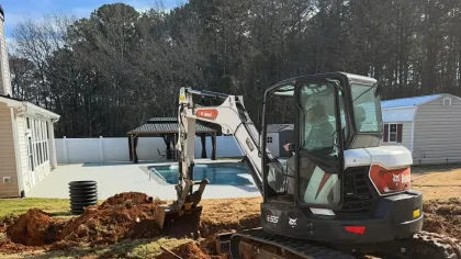 Bobcat compact excavator digging in backyard near pool and house on a sunny day with trees in background.