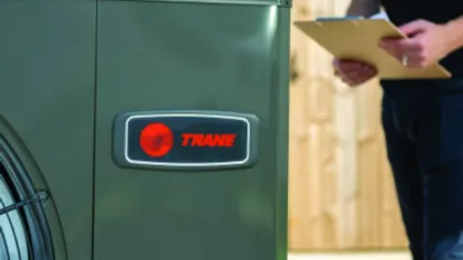 Close-up of a Trane HVAC unit with a technician holding a clipboard in the background near a wooden fence.