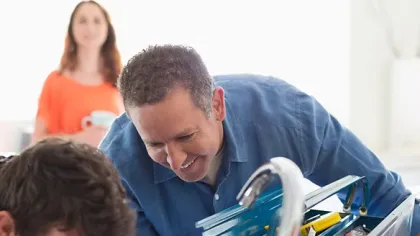 Two men fixing a kitchen sink faucet with a toolbox nearby and a woman watching in the background.