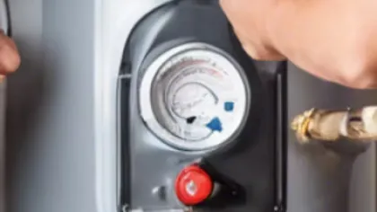 Hands adjusting a pressure gauge on a water heater with red and blue valves and pipes visible.