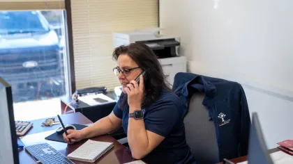 Woman in glasses talking on phone and working at desk with keyboard, notebook, and office printer in background