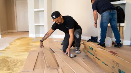 Two men installing wooden flooring in a room with one applying adhesive and the other unpacking materials