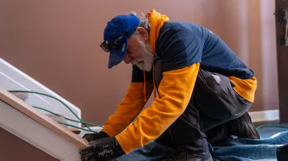 Elderly man in blue cap and orange hoodie installing or repairing wooden stair trim indoors.