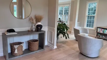 Modern living room with light wood flooring, round mirror, woven baskets, gray chairs, and large windows.